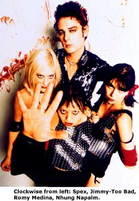 The members of Lolita Storm standing against a white background with bloody red streaks. They are arranged in a diamond formation. A caption at the bottom identifies the members. Jimmy Too-Bad is standing in the back with an aloof look. He has dark, puffy hair, a necklace, and a leather jacket. Spex and Romy Medina are standing in the middle staring at the camera. Spex, on the left, is wearing a shoulderless crop top with elbow cuffs and a choker. Her hair is blonde and straight. Romy, on the right, has a sleeveless vest with a metallic pattern as well as red armbands and a red ribbon in her short, black hair. Nhung Napalm, in front, is wearing a similar vest and is reaching towards the camera with her hand splayed. All four are wearing eyeshadow and the girls all have bright red lipstick.