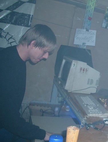 A man sits at a computer holding a mouse on top of a rack module. A CRT monitor and keyboard are visible. The air is smoky.